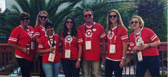 Rachel and six teammates wearing red bullseye jerseys, badges and sunglasses stand against a backdrop of palm trees