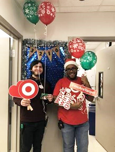 A man in a black shirt stands next to a man in a red shirt and Santa hat holding red and green balloons and assorted work.win.give. signs.