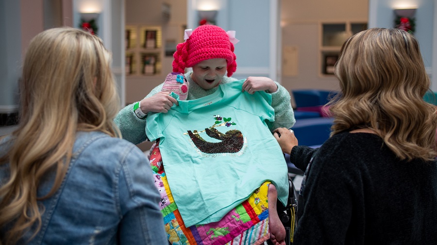 Hallie, wearing a pink beanie, smiles as she holds up a mint green sloth tee she just unwrapped as two designers look on