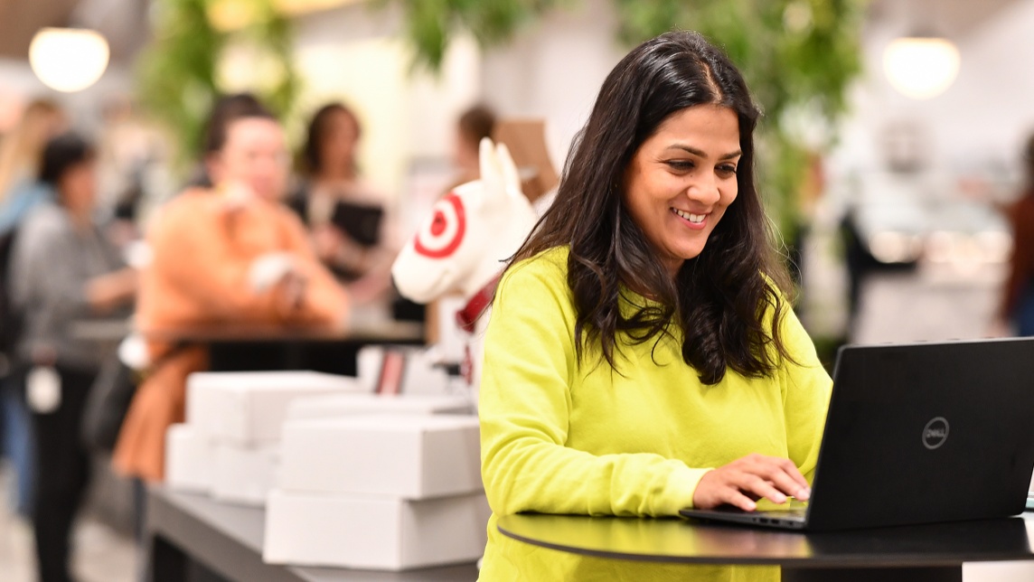 A Target team member in a yellow sweater works on a laptop while smiling.