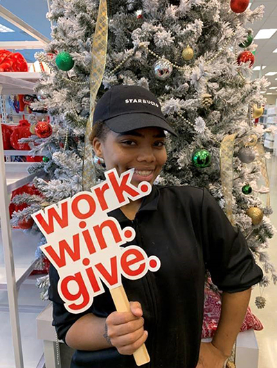 A woman in a black t-shirt and hat that reads Starbucks smiles and holds up a red work.win.give sign in front of a Christmas tree.