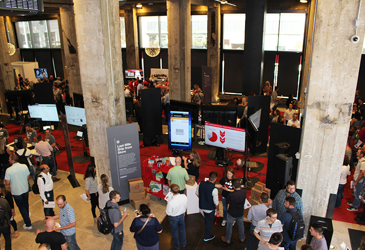 A balcony shot looking down at a large crowd of people milling around, looking at demo booths and talking to team members about their projects.