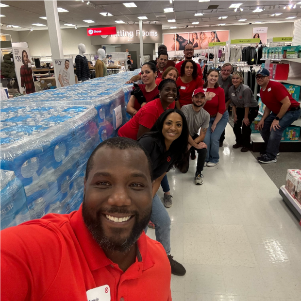 A group of team members smile and pose next to stacks of bottled water in a store.