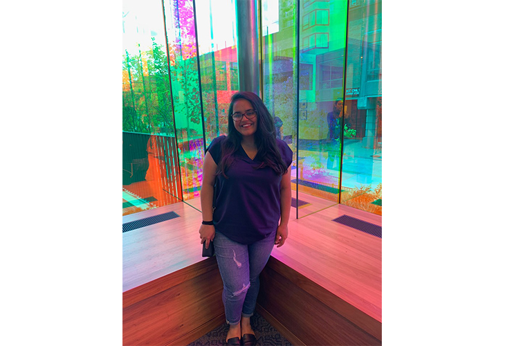 Anushka stands near a multicolored glass window with lights reflecting around her. She's wearing a dark blue top and blue jeans.