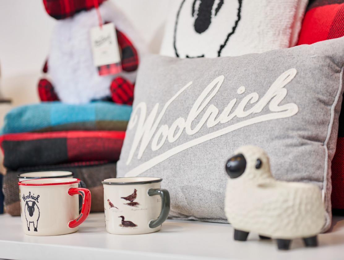 A gray pillow with “Woolrich” written across it and a few Woolrich x Target mugs and a sheep figurine.