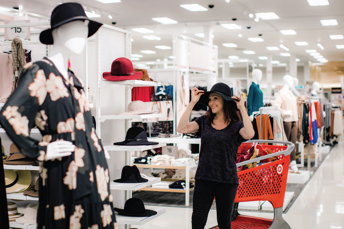 Ellen stands in Target's apparel section trying on a hat next to her red shopping cart filled with products