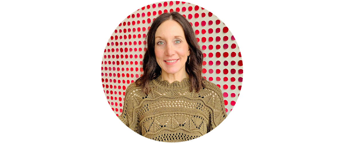 A woman with a green sweater stands in front of a white wall decorated with red circles.