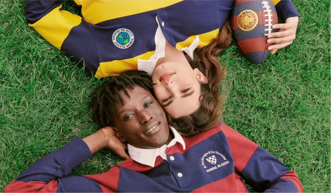 Two models lie side-by-side on the grass wearing striped rugby shirts.