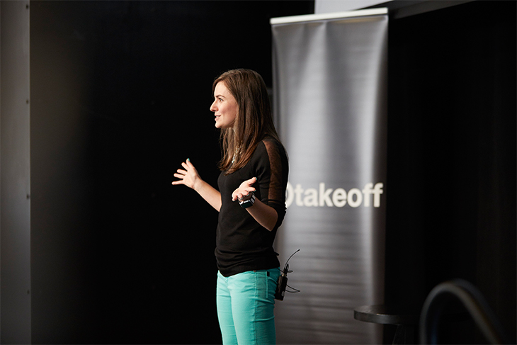 A brunette woman wearing a black top and mint green pants stands onstage with her arms outstretched. Behind her is a black pillar with white text that reads: 'Target Takeoff'