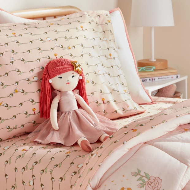 A bed with pink sheets and floral print displays a Princess Ariel plush doll wearing a pink ball gown.