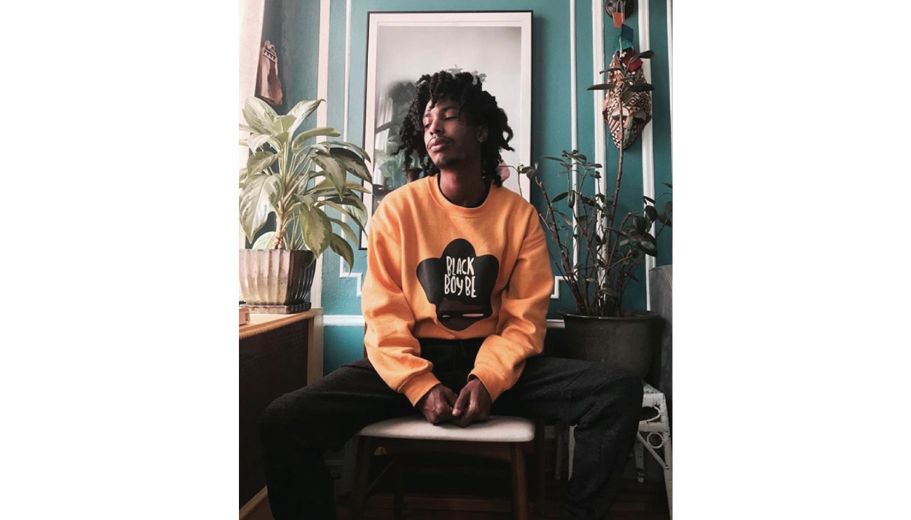 Blu sits against a wall with art and plants, wearing an orange sweatshirt and black pants