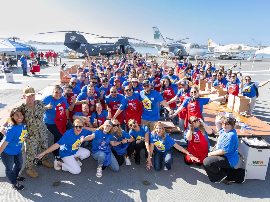 A crowd of people smile at the camera with their arms outstretched. Military planes are in the background, as well as tables for making kitchen essential kits.