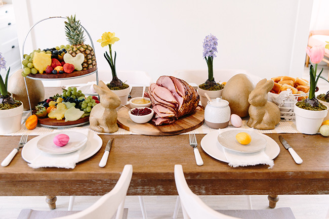 A beautiful tablescape featuring fruit display, dinnerware and napkins, ham and buns, potted flowers and egg and bunny decor
