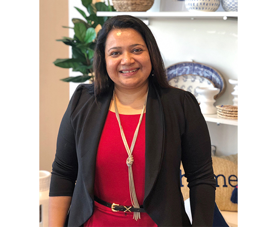 Sanhita stands in front of a shelf of Home products. She's wearing a red dress, gold necklace and black blazer.