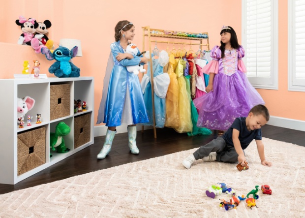 Two girls dresses as Disney Princesses play near dress rack and other Disney Toys while boy plays with Disney toys on the floor in a kids room.
