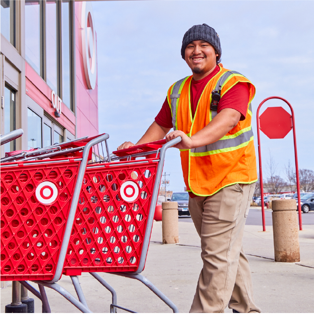 A Target team member in an orange and yellow safety vest smiles while pushing red Target carts into a store.