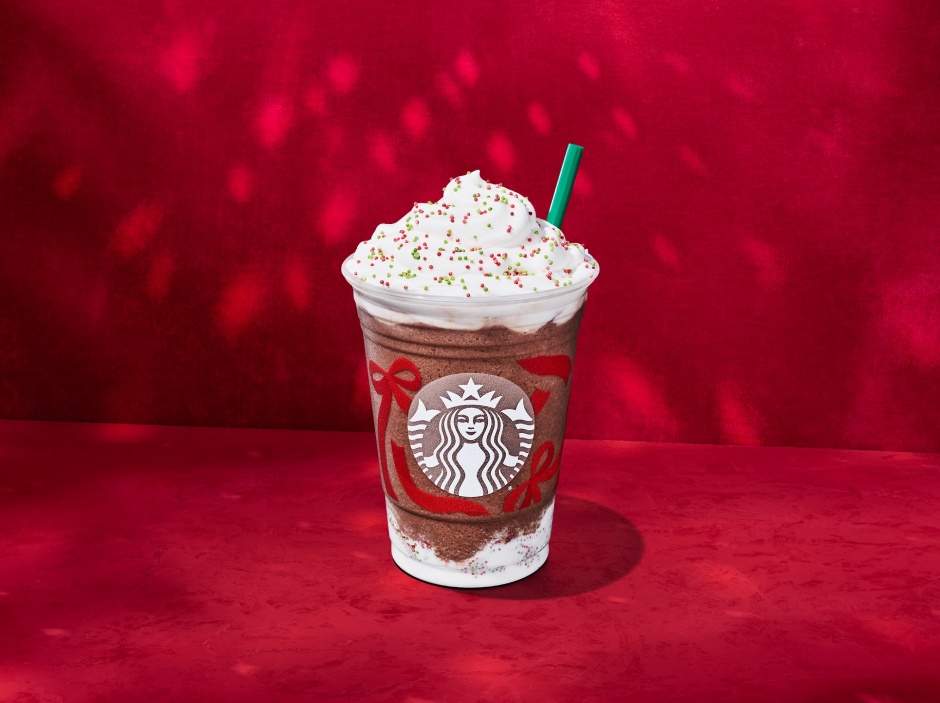 A Starbucks Frozen Peppermint Hot Chocolate topped with whipped cream and red and green sprinkles, in a clear holiday cup with red ribbon designs, set against a red background.