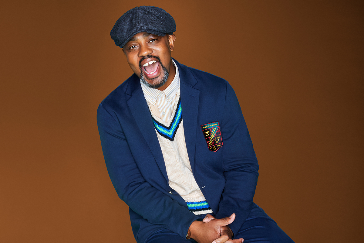 A picture of Houston White sitting down wearing a cap, navy blue blazer, a sweater and navy blue pants from his collection.