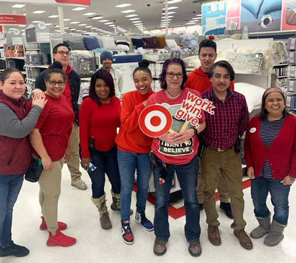 A group of 10 team members in red shirts stand in a Target bedding aisle laughing and holding assorted signs. Cymon stands in front; she has purple hair and glasses and is in a red shirt and blue jeans.