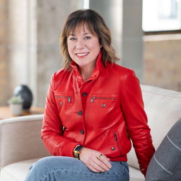 Adrienne Costanzo smiling, sitting on a sofa at Target headquarters. She’s wearing a red jacket, blue jeans and a watch.