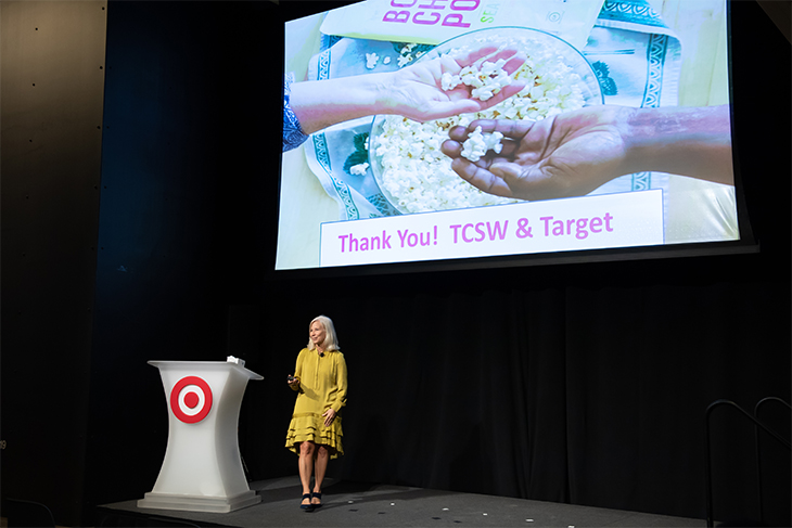 Angie stands onstage in a yellow dress in front of a screen with a photo of hands in a popcorn bowl. Text reads Thank You! TCSW and Target