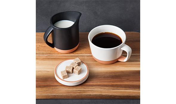 A white coffee mug filled with coffee next to a black pitcher of cream and plate of sugar cubes on a wooden tray