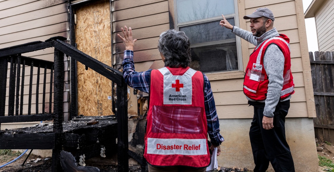 Two adults in Red Cross vests survey a fire damaged house.
