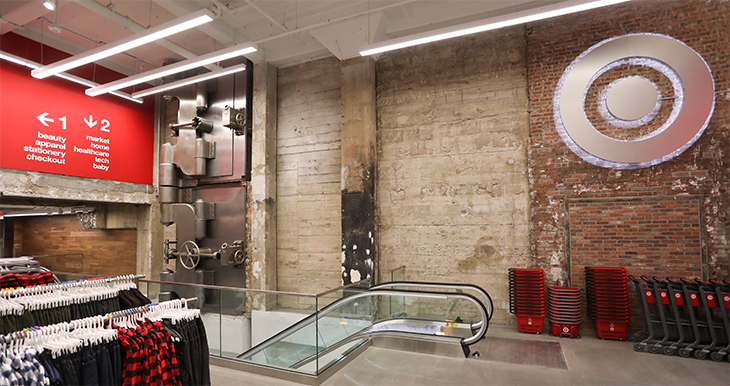 The entry of a Target store with a brick wall, escalator, red signs, baskets and apparel racks.