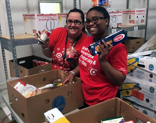 Two team members in red shirts stand in the backroom of a food bank sorting cardboard boxes of food donations.