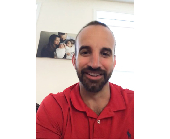 Ryan smiles at the camera, wearing a red shirt in front of a family photo on the wall