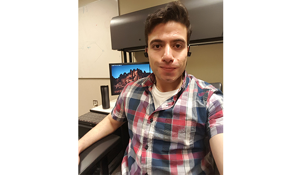 Asad sits at his home office with his computer. He has short brown hair and is wearing a plaid shirt and ear buds.