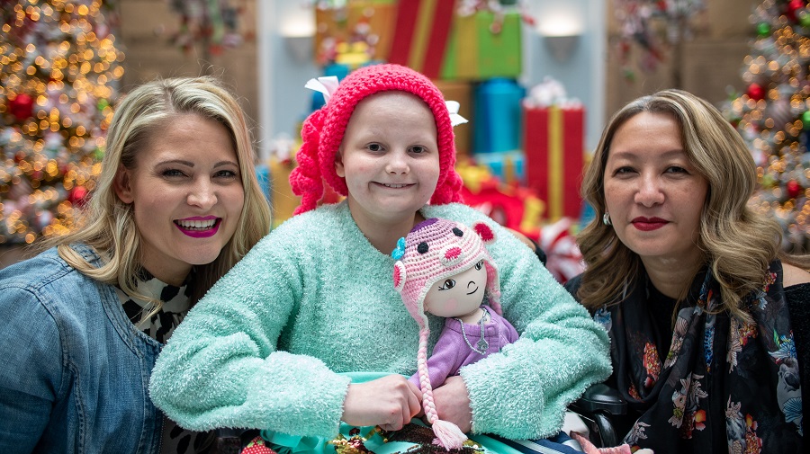 A head-and-shoulders shot of Hallie between two designers. Hallie is holding the new doll they gave her and smiling.