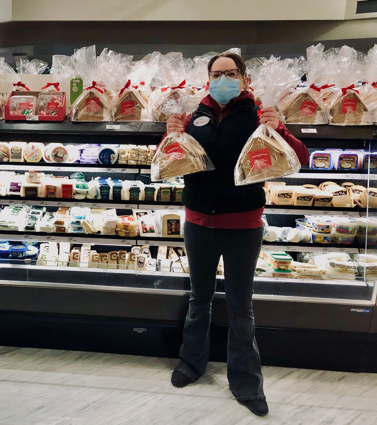 Mariah, wearing a vest, name tag and face mask, holds a gingerbread house in front of a cooler of cheeses