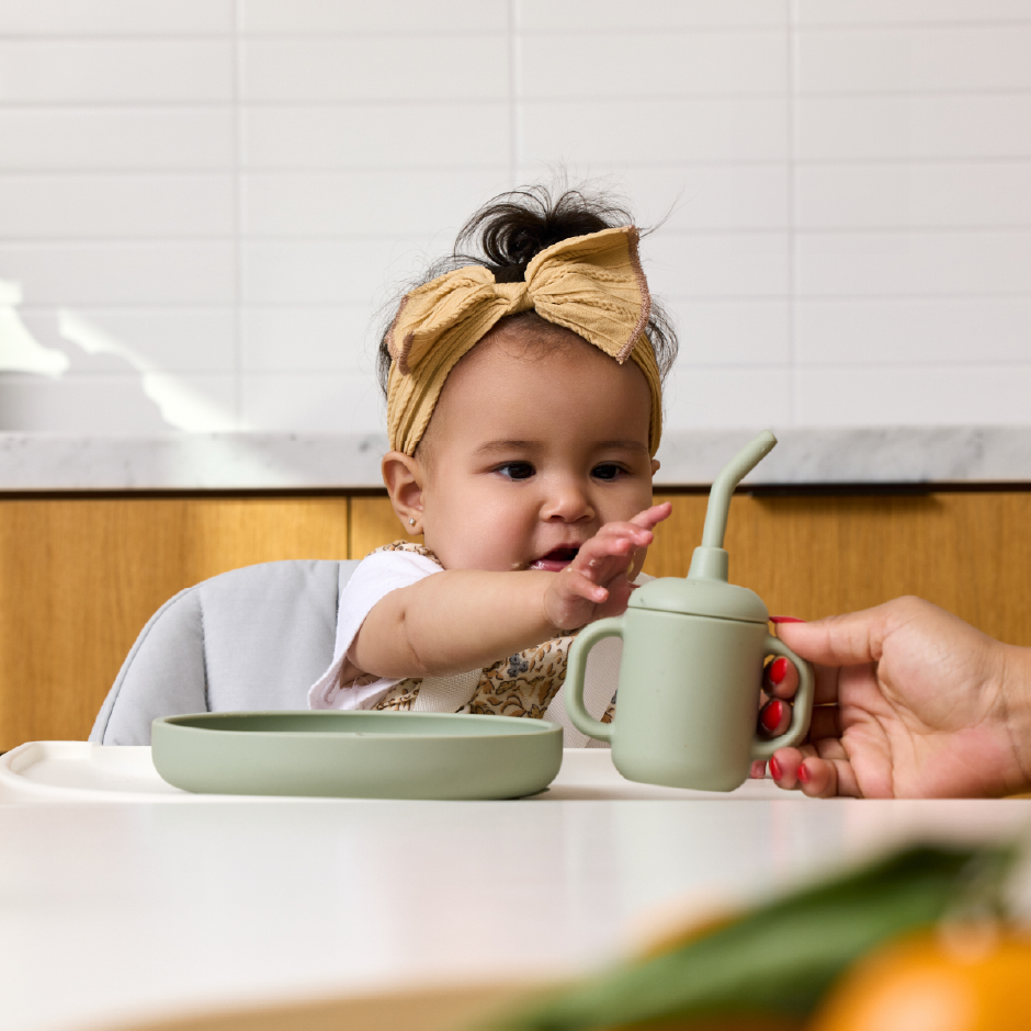 A baby with a bow in her hair reaches for a sage green cup being handed to her.