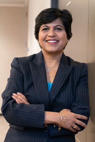 A smiling woman stands in her office with arms crossed. She's wearing a striped navy blazer and blue shirt.