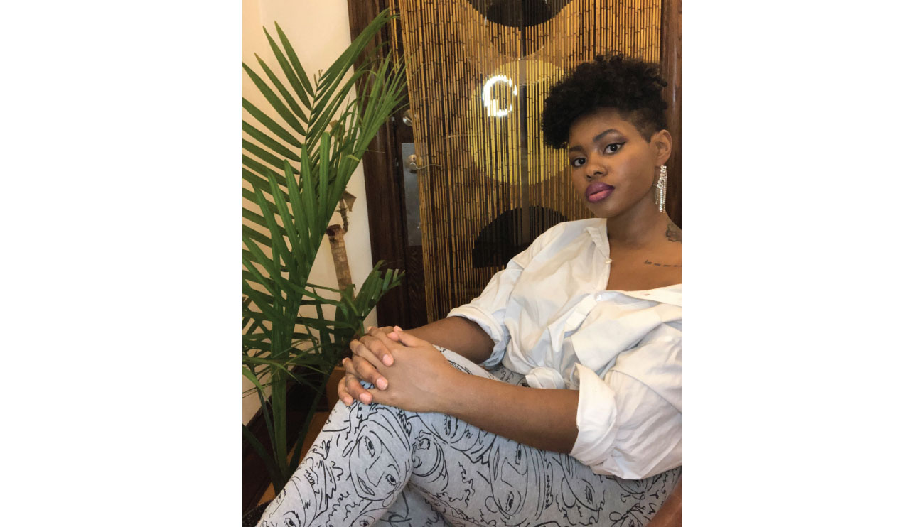 Shanetta sits against a door next to a potted plant wearing a white top, leggings with illustrated faces and long, sparkly earrings
