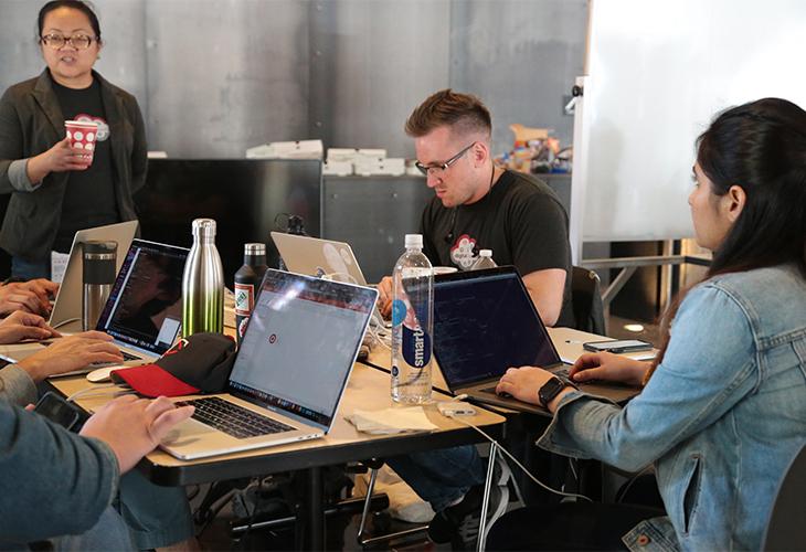 A man and a woman sit at a long table coding on their laptops while another woman watches, holding a cup. The hands of other team members and their laptops are visible on the other side of the table.