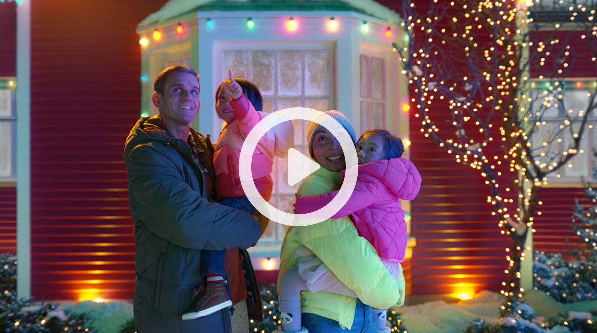 Standing outside in front of Christmas lights, a man holds a young girl wearing a pink coat, next to him a woman holds another young girl wearing a pink coat