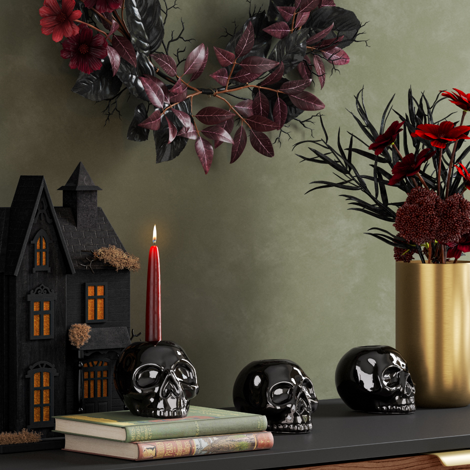A Halloween display of skull candleholders, a black house and a gold vase with dark flowers.