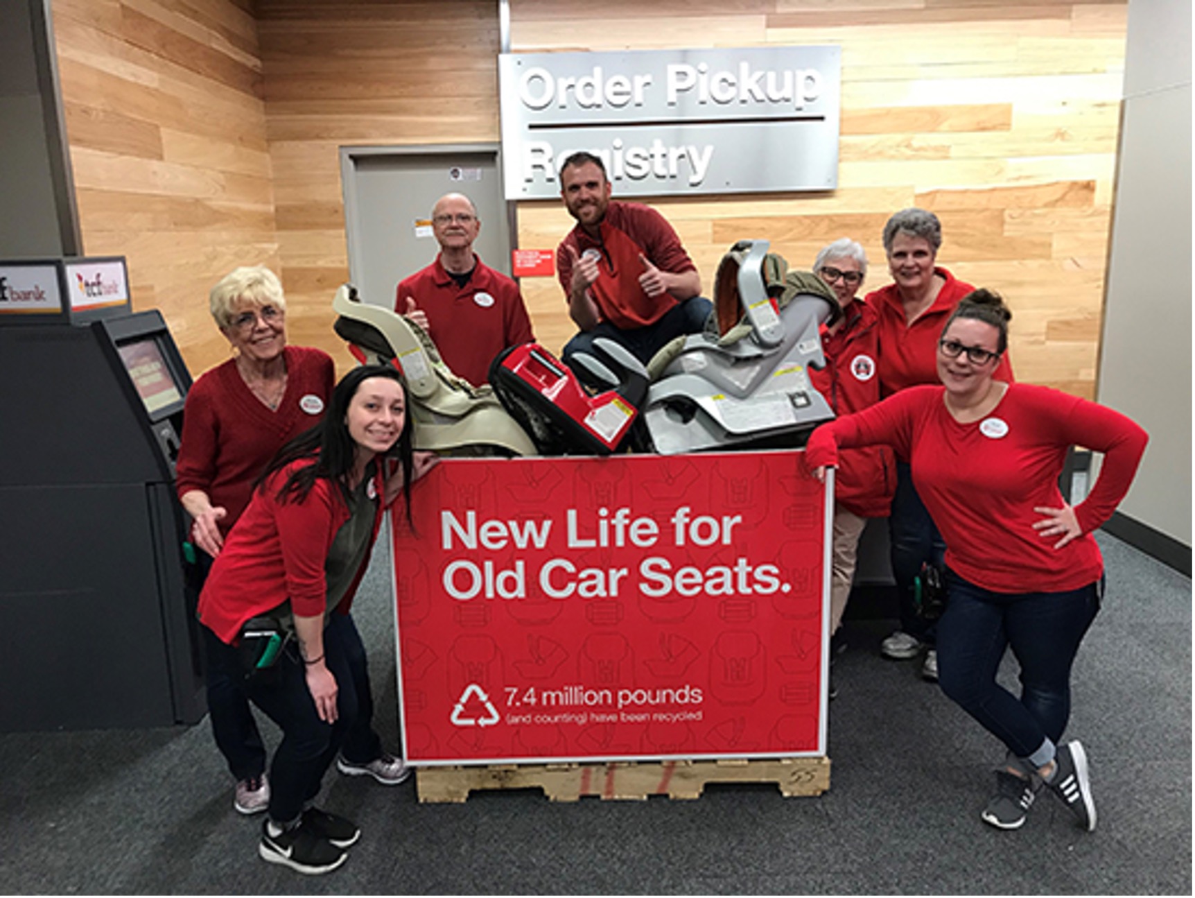 A photo of seven team members in red shirts, jeans and name badges, smiling as they gather around a large drop-off box at their store filled with used car seats for recycling.