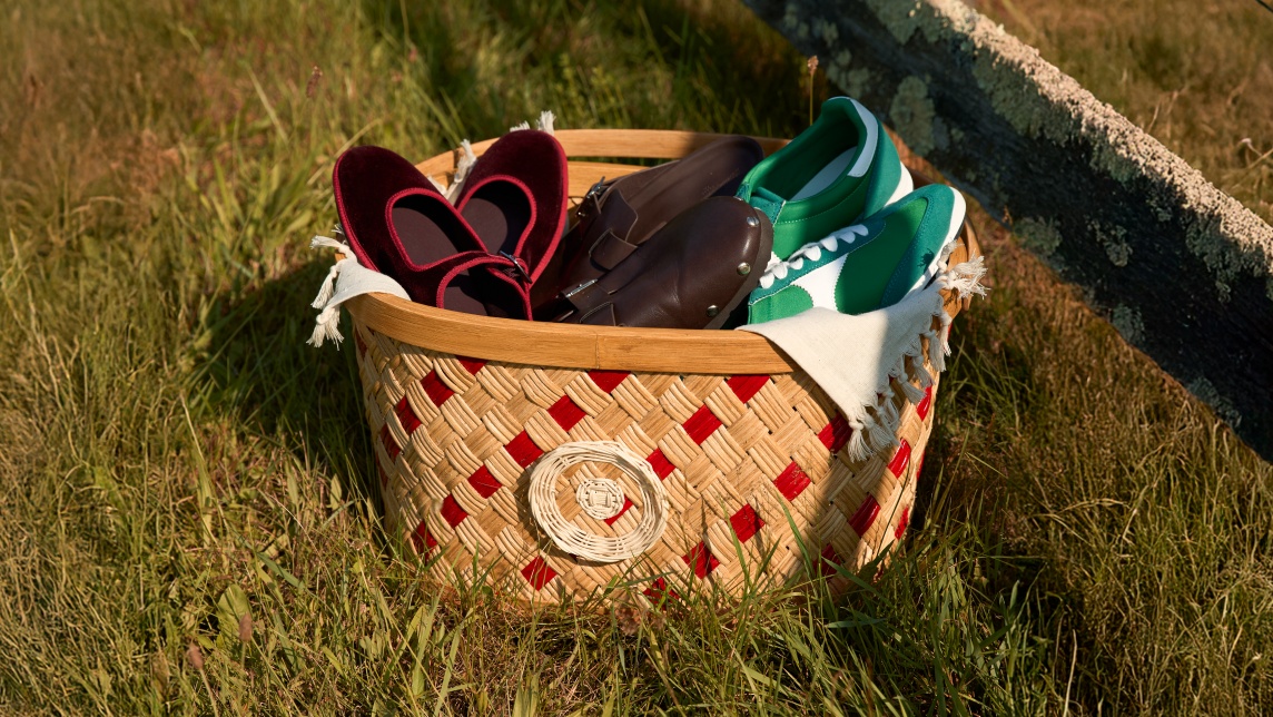A woven red-and-tan basket sits on grass, filled with three pairs of shoes: red velvet flats, brown loafers, and green-and-white sneakers, with a cream cloth draped over the side.