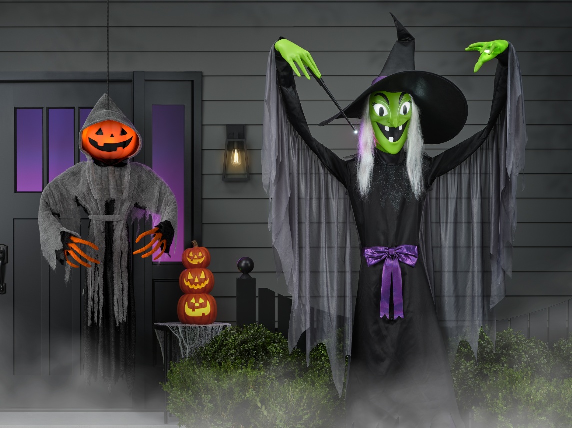 A Halloween yard display features a pumpkin-headed figure in a hooded cloak and a tall green-faced witch with a pointed hat and flowing sleeves, posed near stacked jack-o’-lanterns in front of a house.
