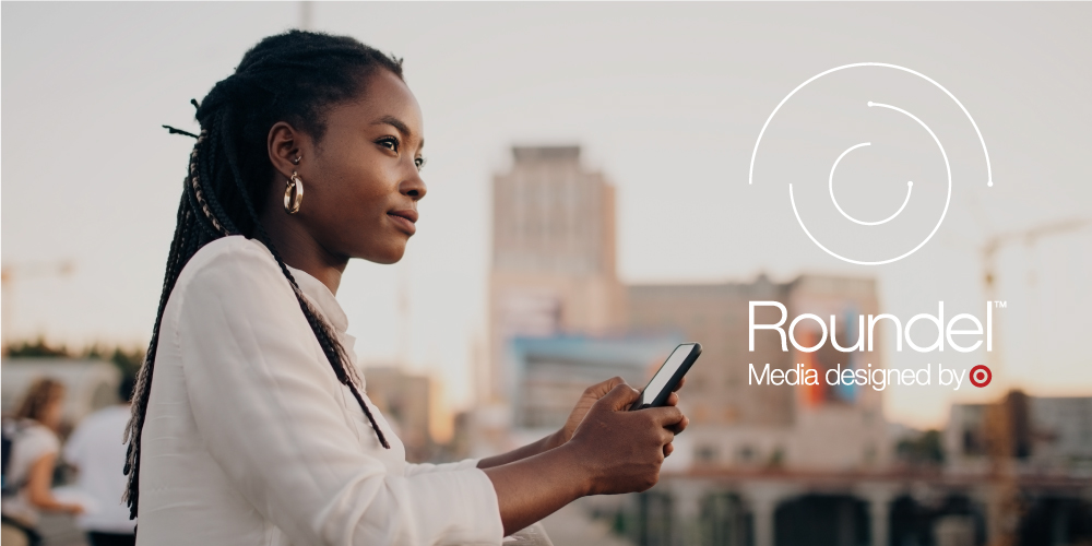 A woman in a white shirt holds a phone in front of a cityscape; she is next to a deconstructed Target symbol with the words "Roundel Media designed by Target."