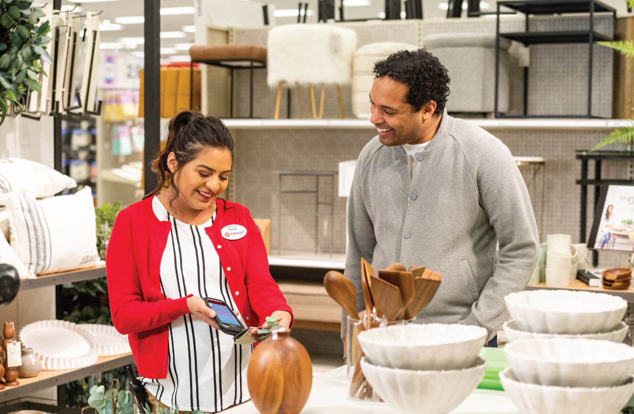 A team member in a red shirt shows a guest in a gray shirt her smartphone screen as she assists him in the Home decor department. They're surrounded by furniture and products.