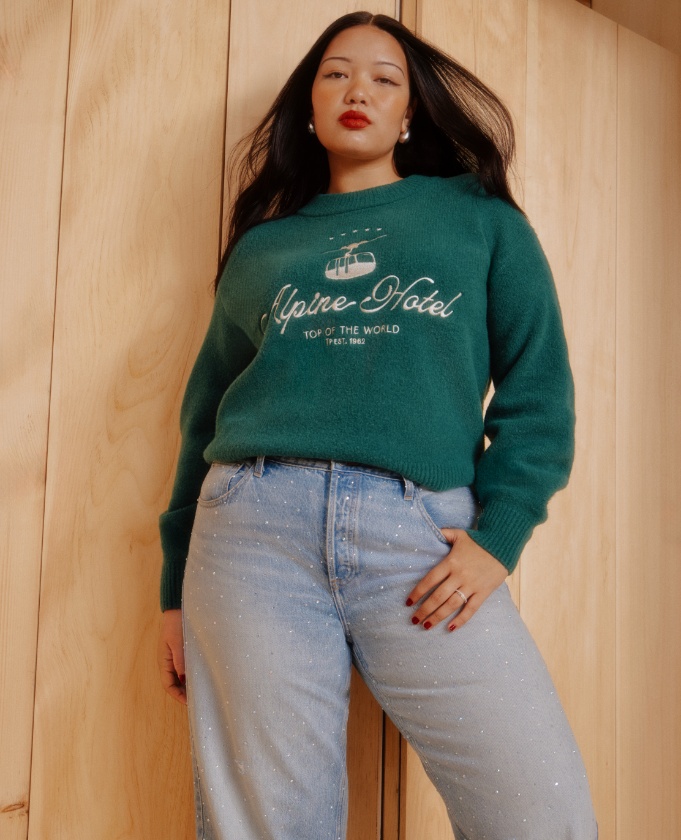 a model wearing a green sweater that says “Alpine Hotel” in script