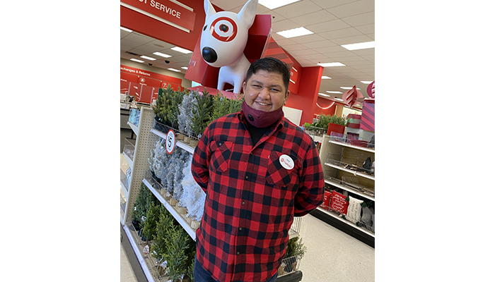 Jose stands at the entrance near a statue of Bullseye the dog. He's wearing a red plaid shirt and mask.