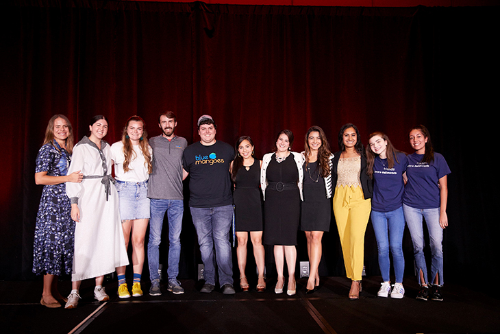 The 11 members of the first cohort stand together on the stage with a red curtain after their presentations