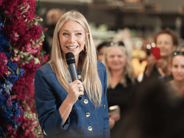Gwyneth Paltrow smiling and holding a microphone in front of a floral backdrop and group of onlookers.