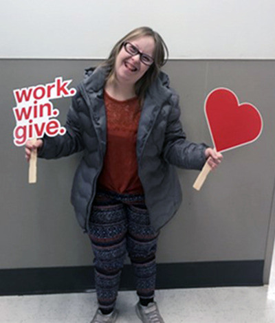 A woman wearing glasses, a gray jacket, rust shirt and patterned leggings smiles as she holds up a work.win.give. sign and another heart-shaped sign.