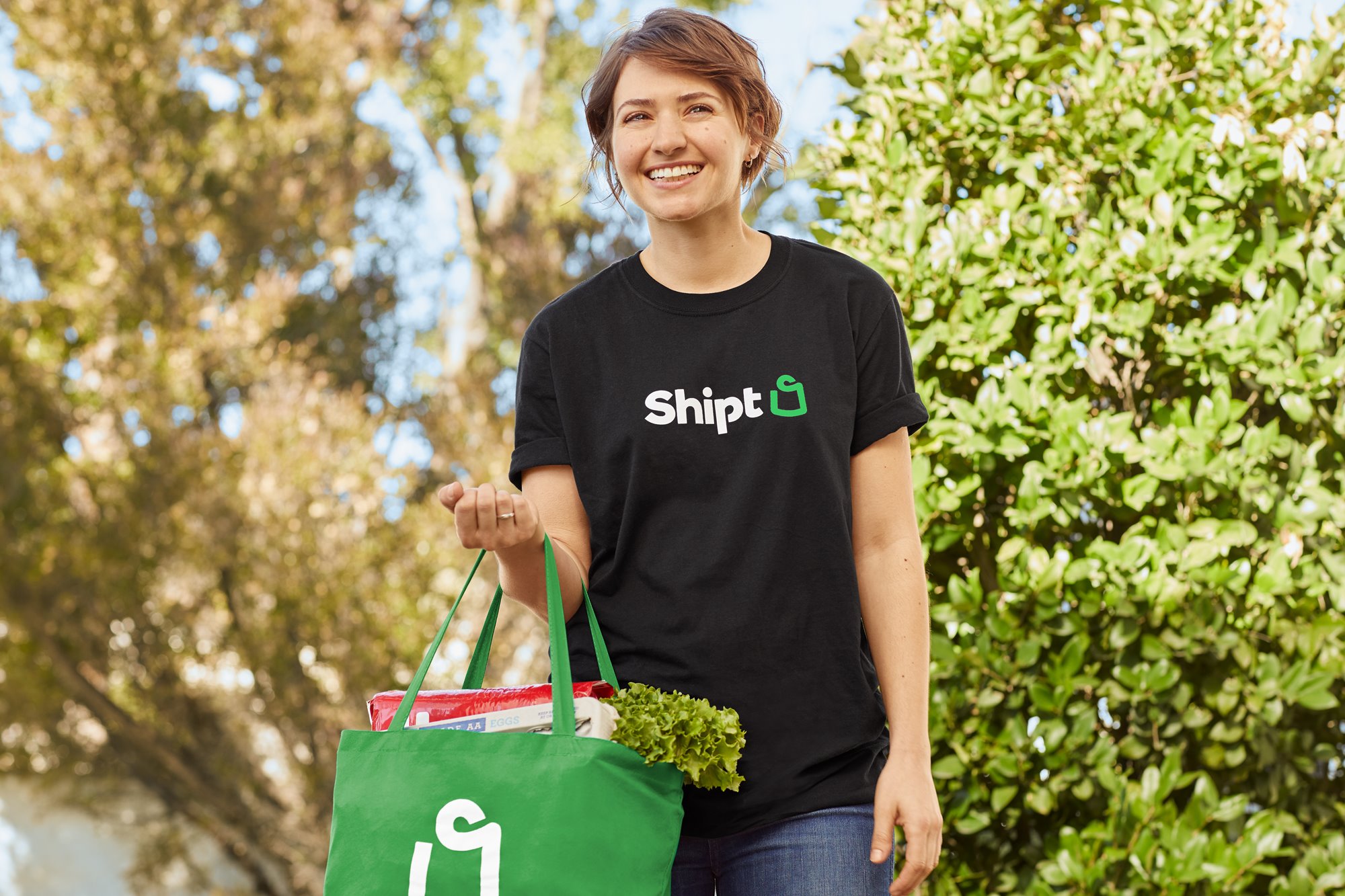 A Shipt Shopper in a black t-shirt smiles as the holds a green bag of groceries on her arm.